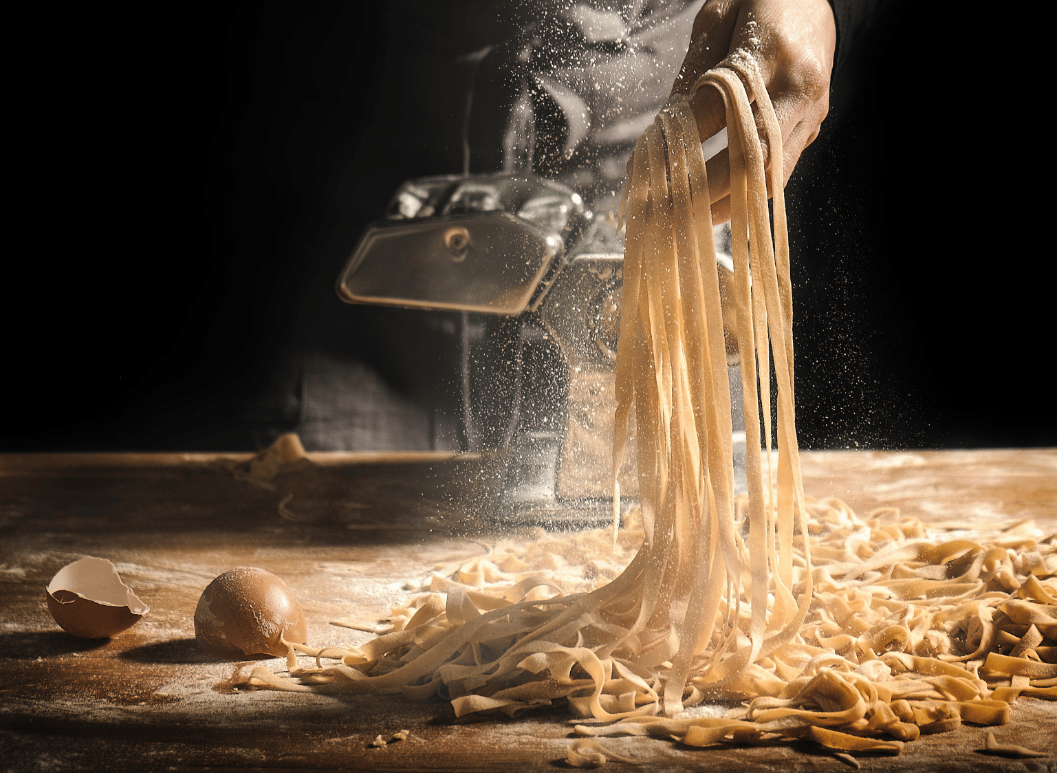 A person making fresh pasta by hand, holding a bunch of long pasta with a pasta machine in the background, on a floured wooden surface with eggs and pasta scraps.