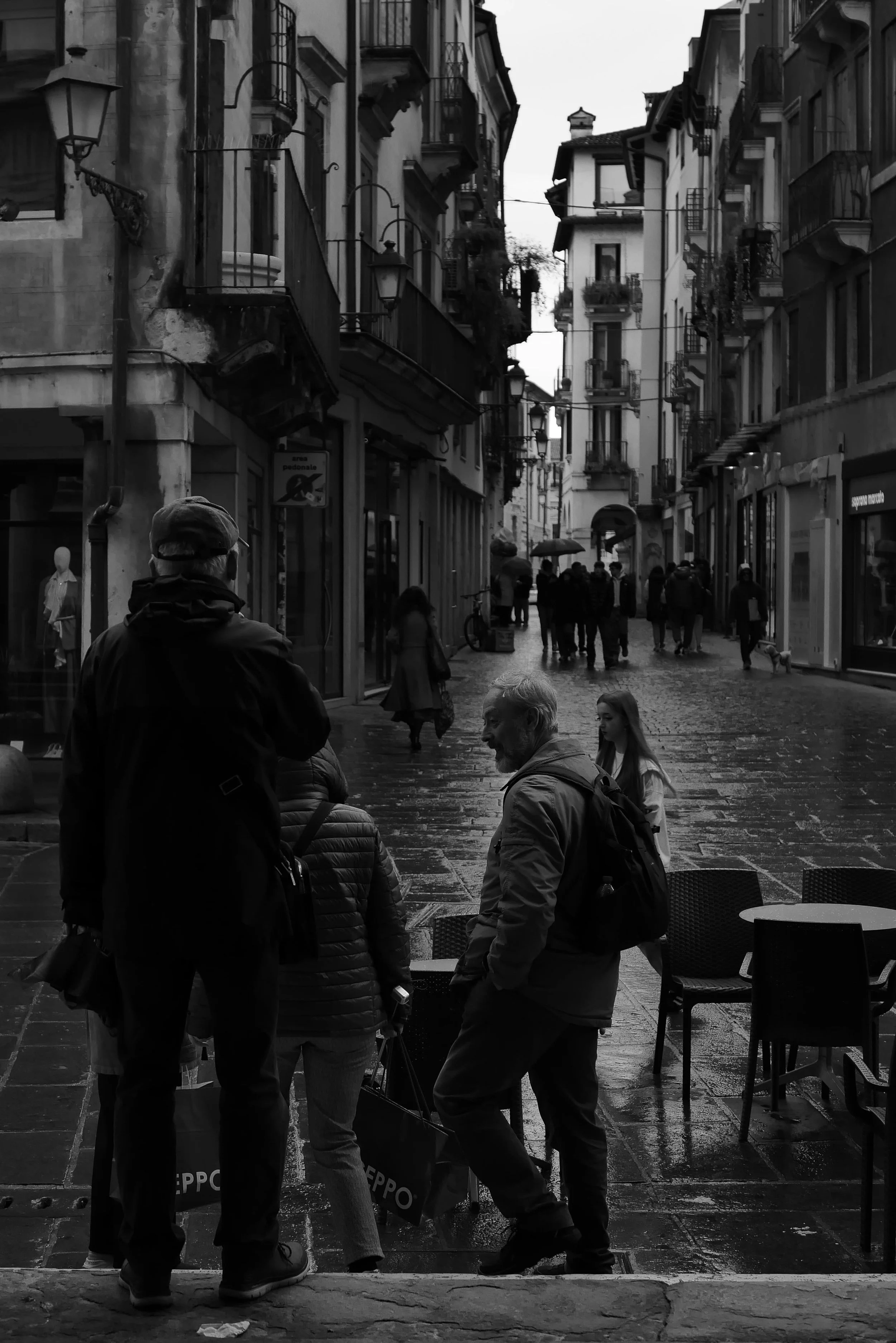 A black and white photo of a European city street with buildings on both sides. Several people walk and stand around, some holding umbrellas. The street is wet, possibly from rain, and there are outdoor seating areas with chairs and tables.