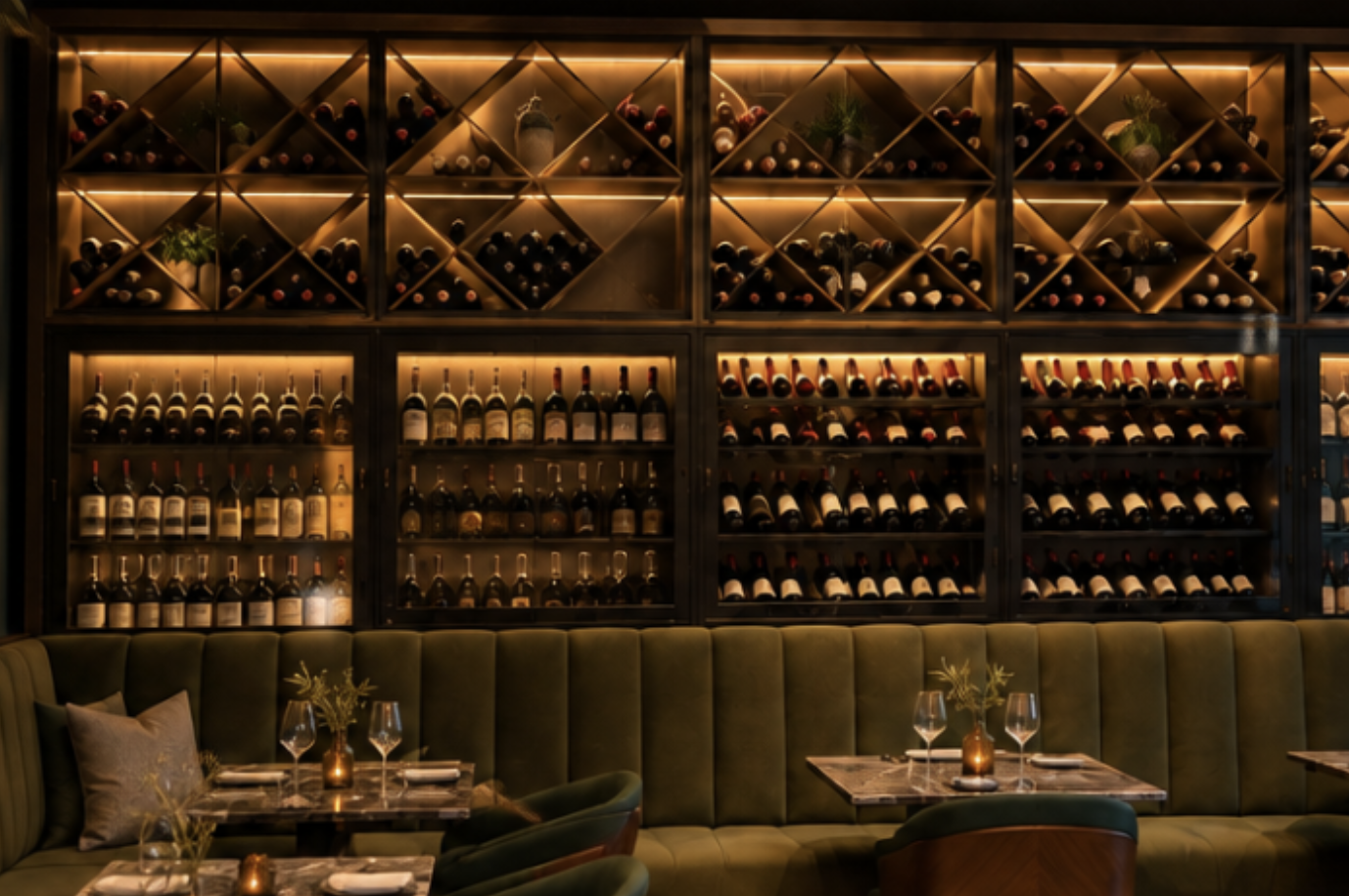Wine bottles displayed on wooden shelves with cabinets in a dimly lit restaurant or bar, with tables set with glasses and small potted plants in the foreground.