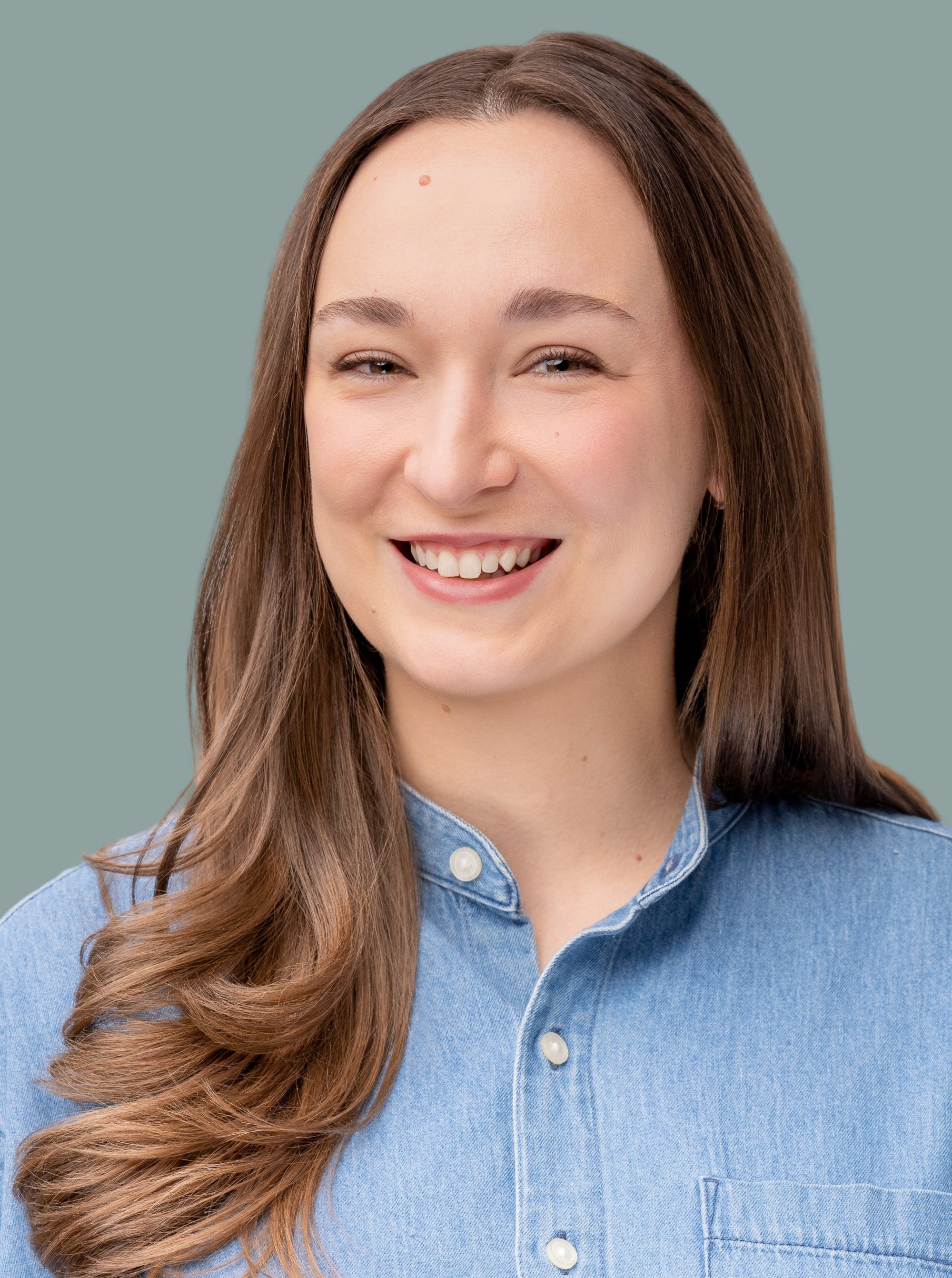 Close-up of a woman with long, wavy brown hair, smiling, wearing a denim shirt, against a plain background.