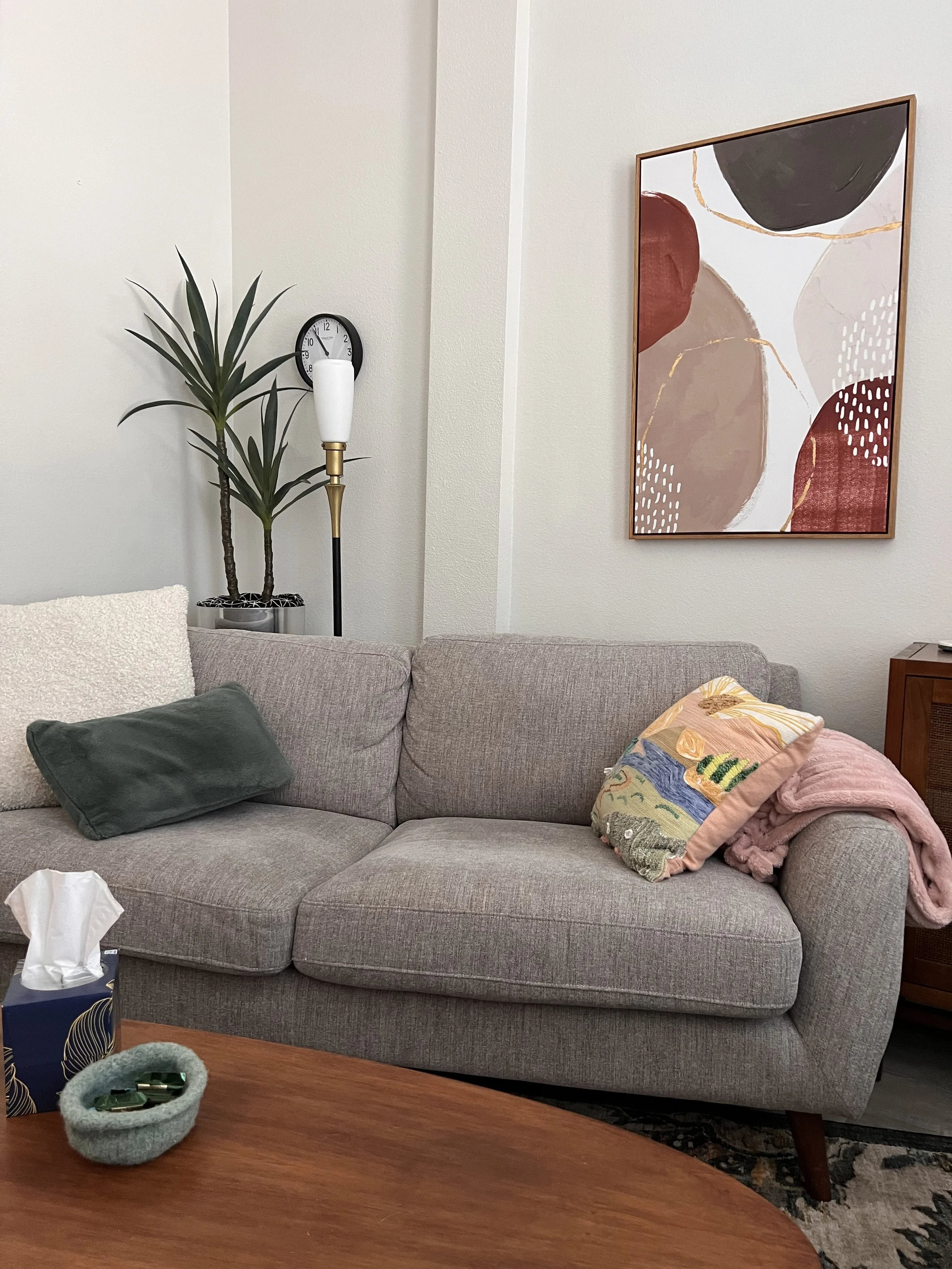 A gray sofa with a white textured pillow, a green pillow, and a colorful embroidered pillow. On the sofa, there is a pink blanket. Behind the sofa, there is a wall with a framed abstract painting and a tall potted plant. A clock is mounted on the wall between the plant and the painting. A floor lamp with a white shade is also in the corner.