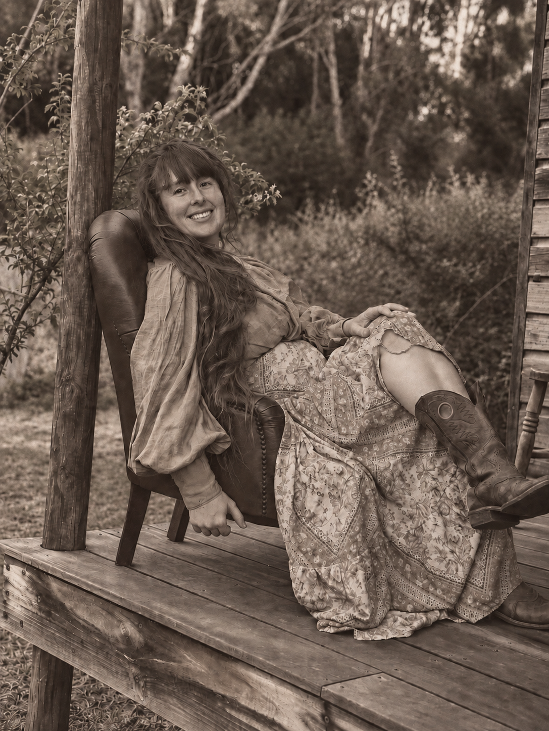 A woman with long hair sitting on a wooden porch, smiling at the camera, outdoors with trees and bushes in the background, sepia tone.