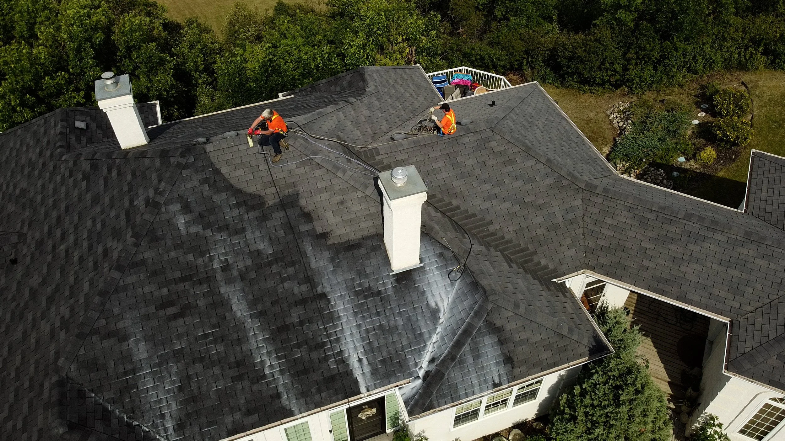 Aerial view of two workers cleaning or repairing a dark gray shingle roof of a large house, with trees surrounding the property.