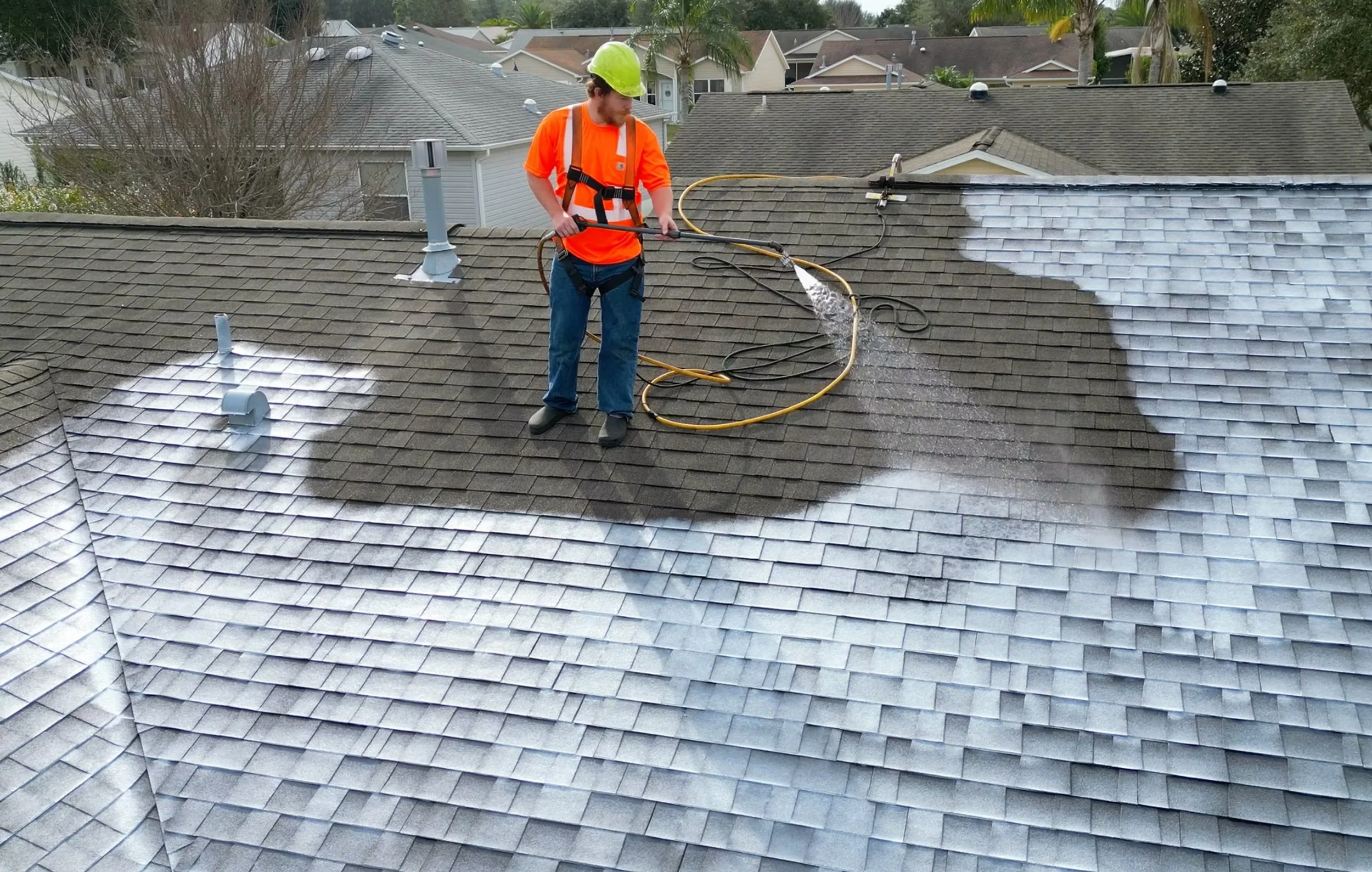 A man wearing safety gear pressure washing a roof with a pressure washer on a residential house.