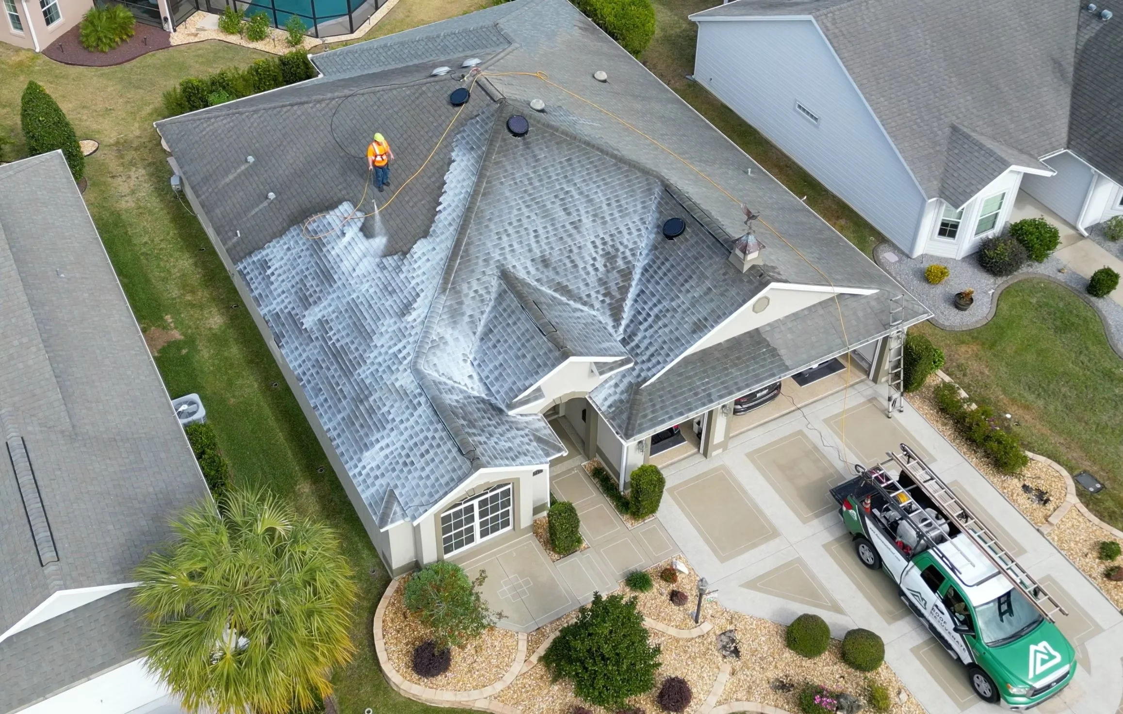 An aerial view of a house's roof during cleaning, with a worker in an orange safety vest and yellow helmet using a hose. The house has a grey shingle roof and is surrounded by a landscaped yard with bushes, trees, and a light-colored concrete driveway. A large window and a vent are visible on the house, and there is a green vehicle parked nearby.