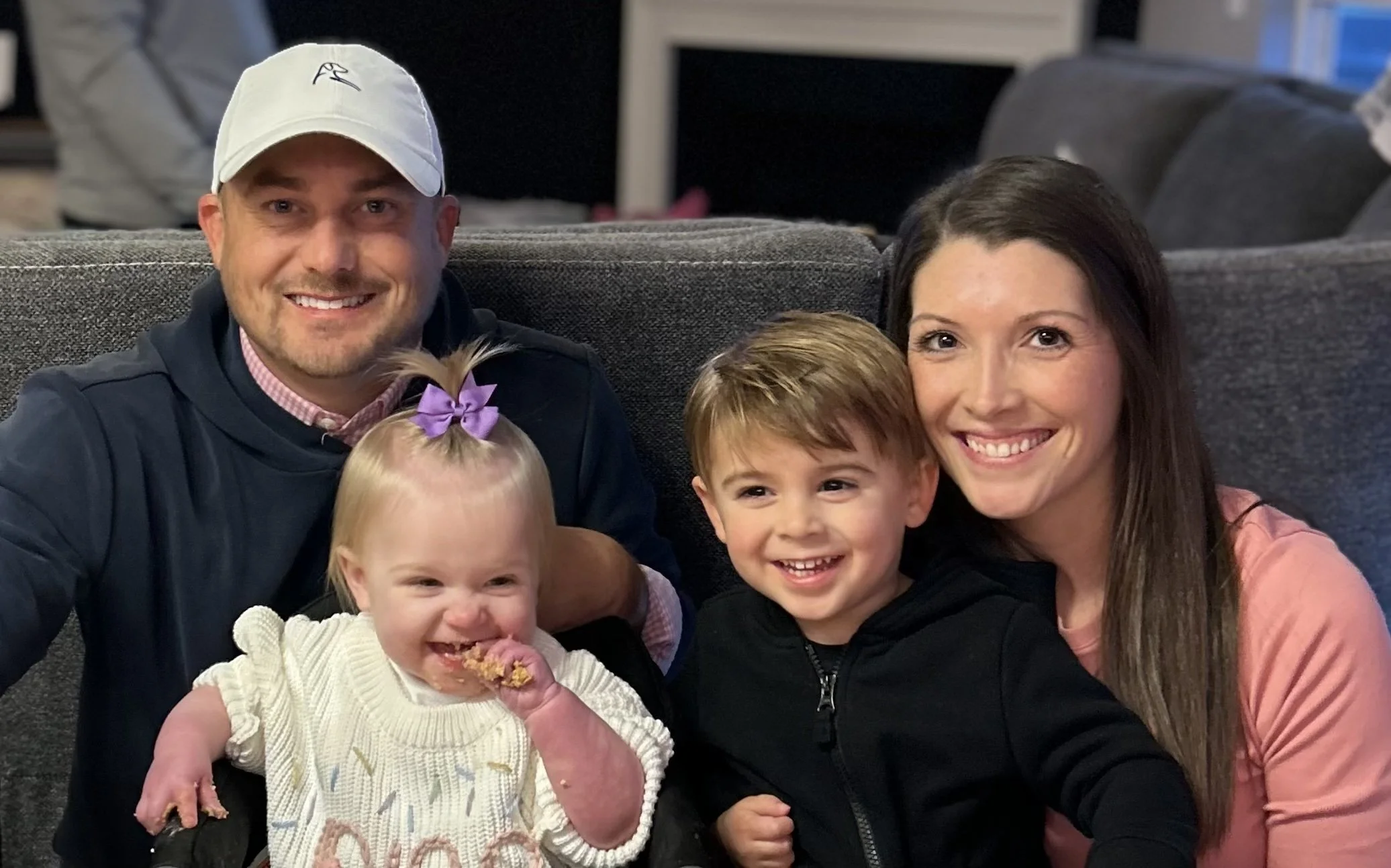 A family of five, including two young children, is gathered together on a couch, smiling at the camera. The man is wearing a white cap and a dark jacket, the woman has long brown hair, and the children have light brown hair. The girl is eating a cookie, and the boy is smiling.