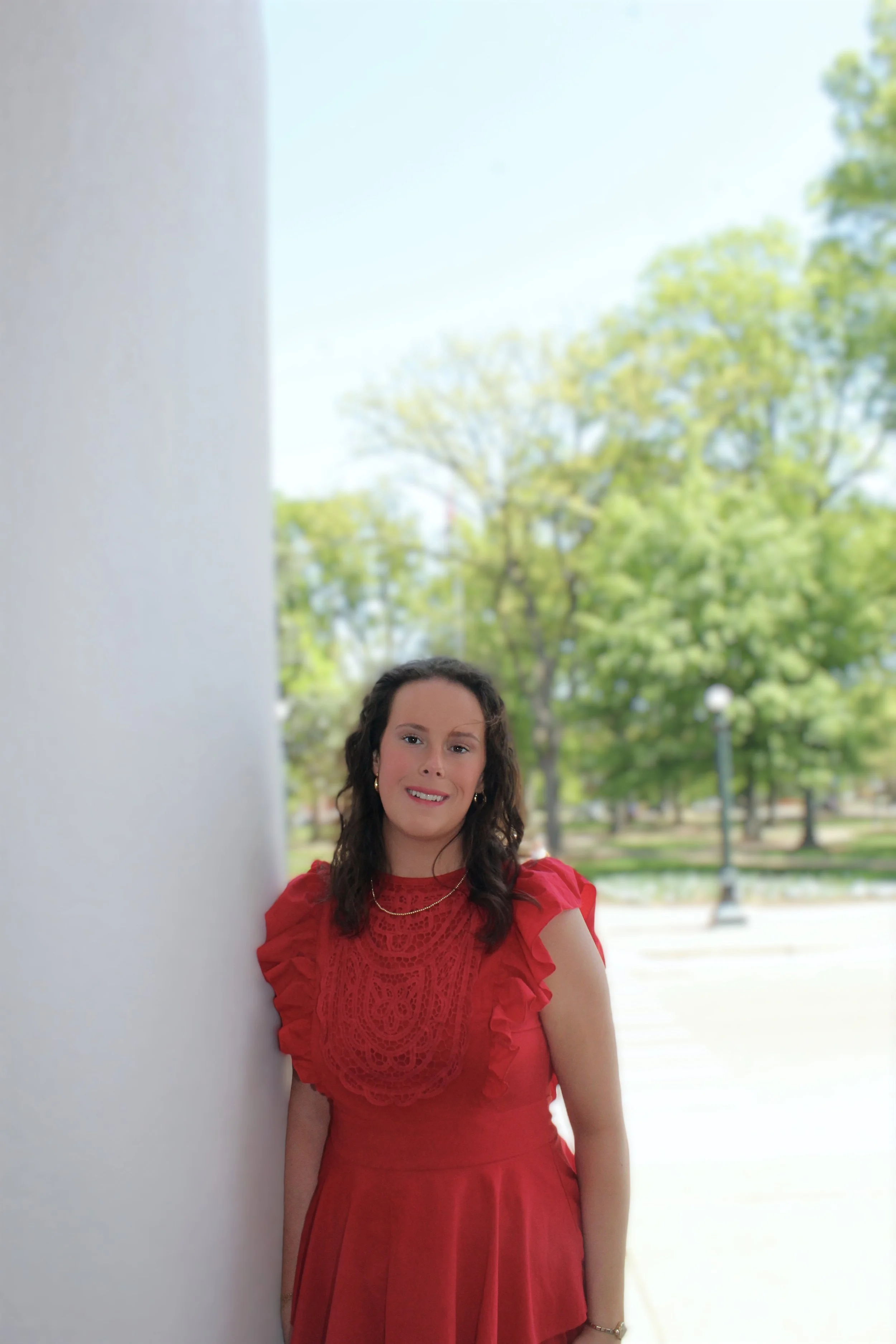 A woman in a red dress standing outdoors, smiling, with trees and a park in the background.