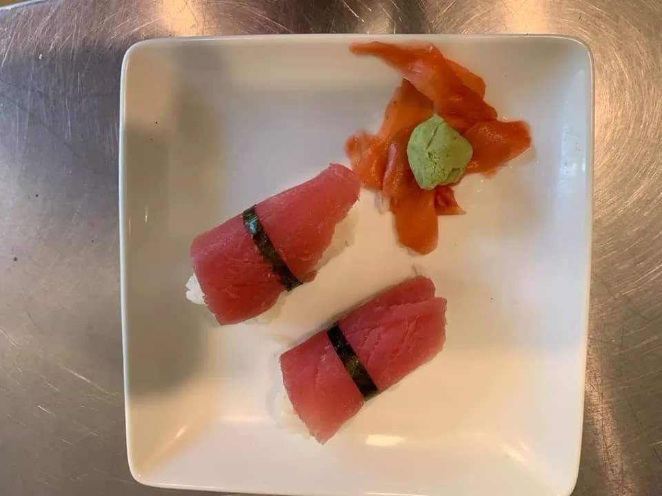 Two pieces of tuna nigiri with seaweed strips, a small serving of ginger, and a dollop of wasabi on a white square plate.