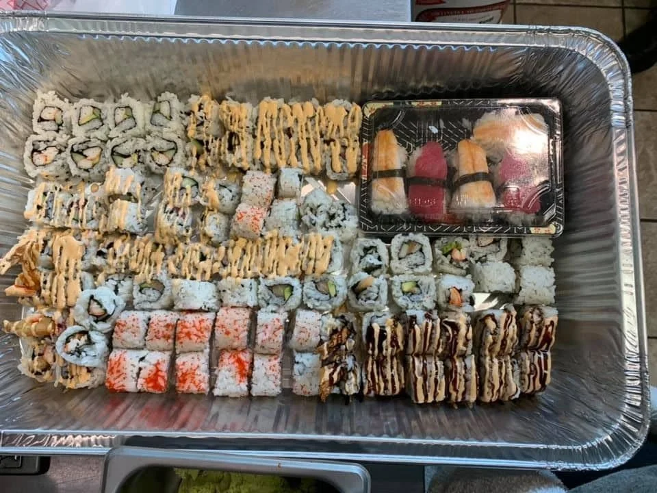 A large tray of assorted sushi rolls, including several types of maki and nigiri, with a plastic container of extra sushi on the side.
