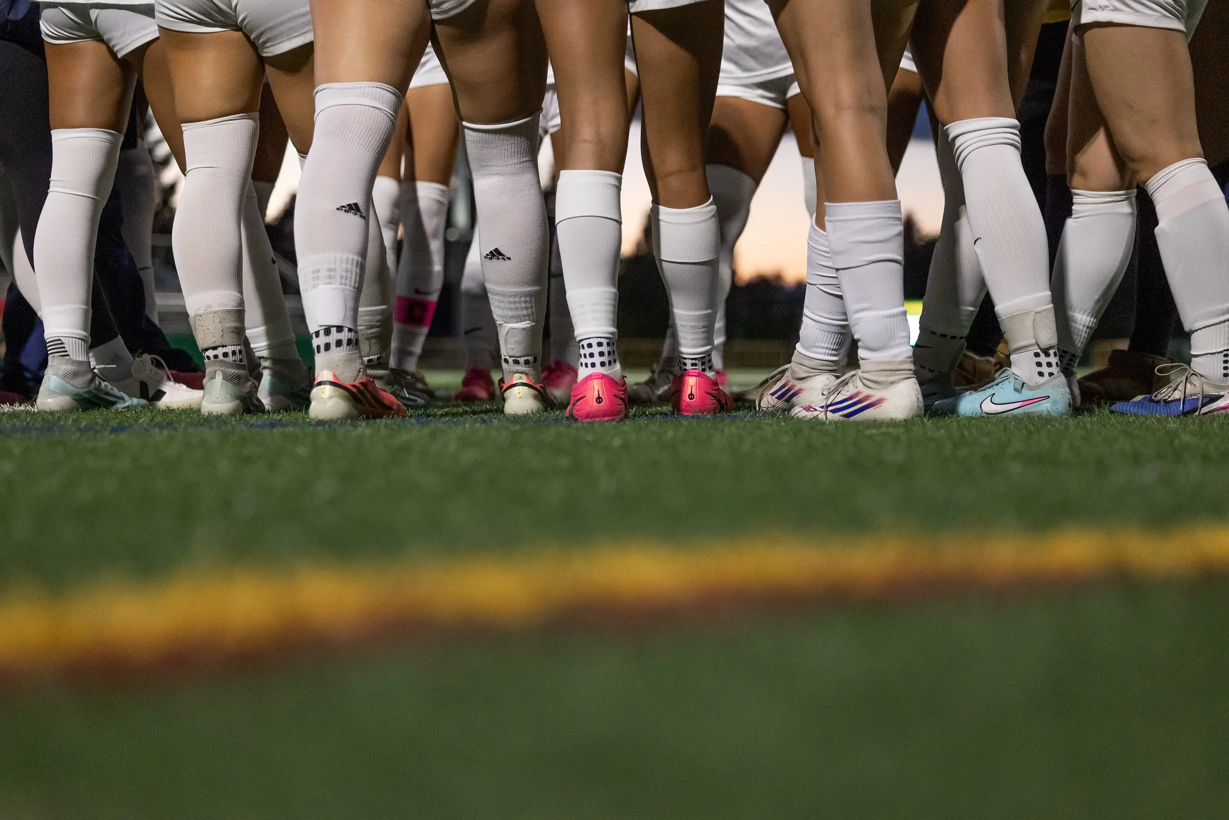 Close-up of soccer players' legs and feet, wearing white socks and cleats, lined up on the field during sunset.