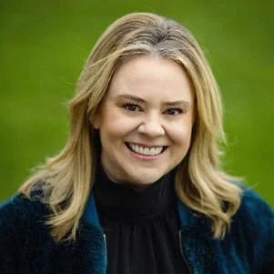 A smiling woman with blonde hair, wearing a black top and a dark blue jacket, outdoors on a green background.