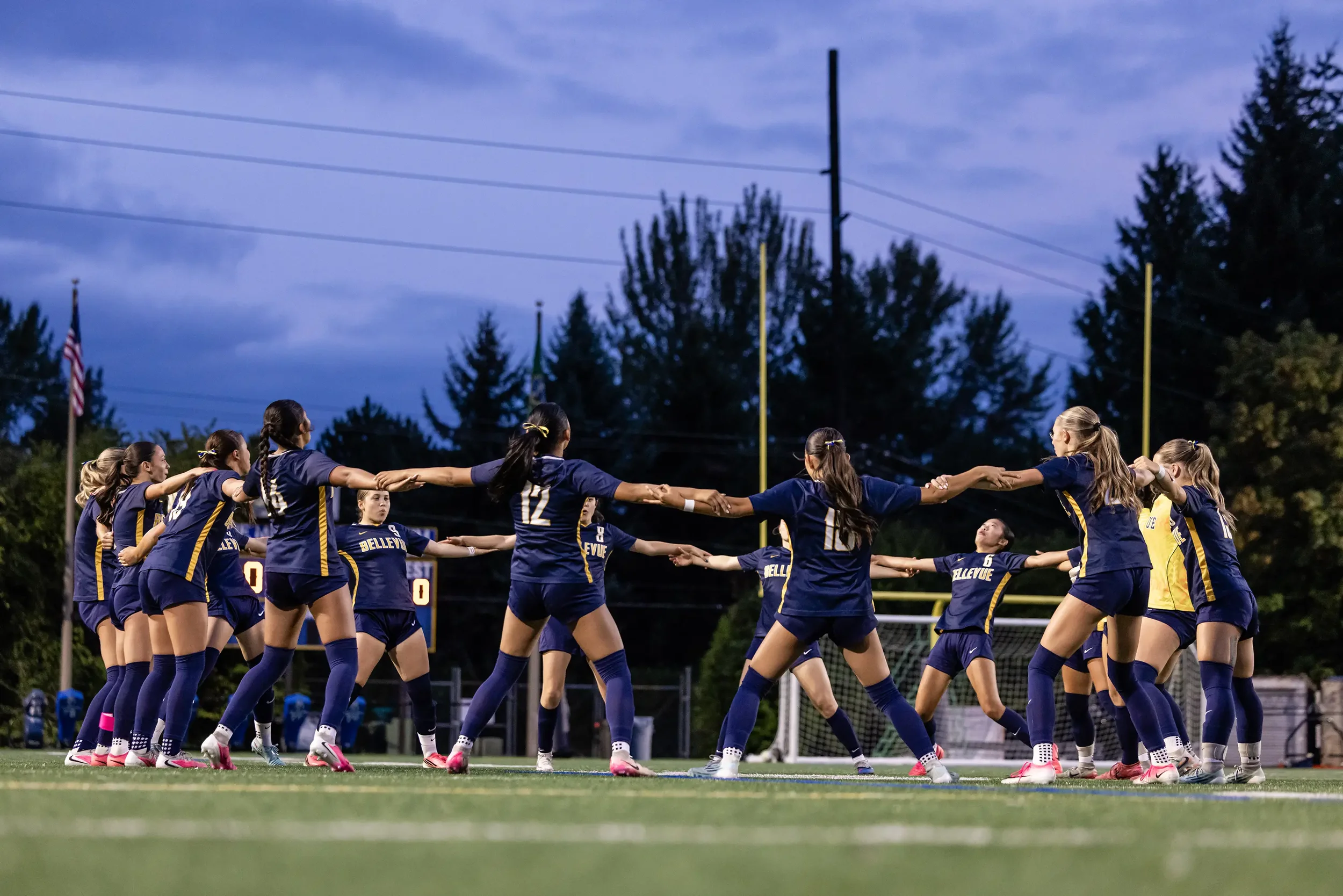 Young female soccer players in blue and yellow uniforms forming a line and holding hands on a football field during dusk, with trees and flags in the background.
