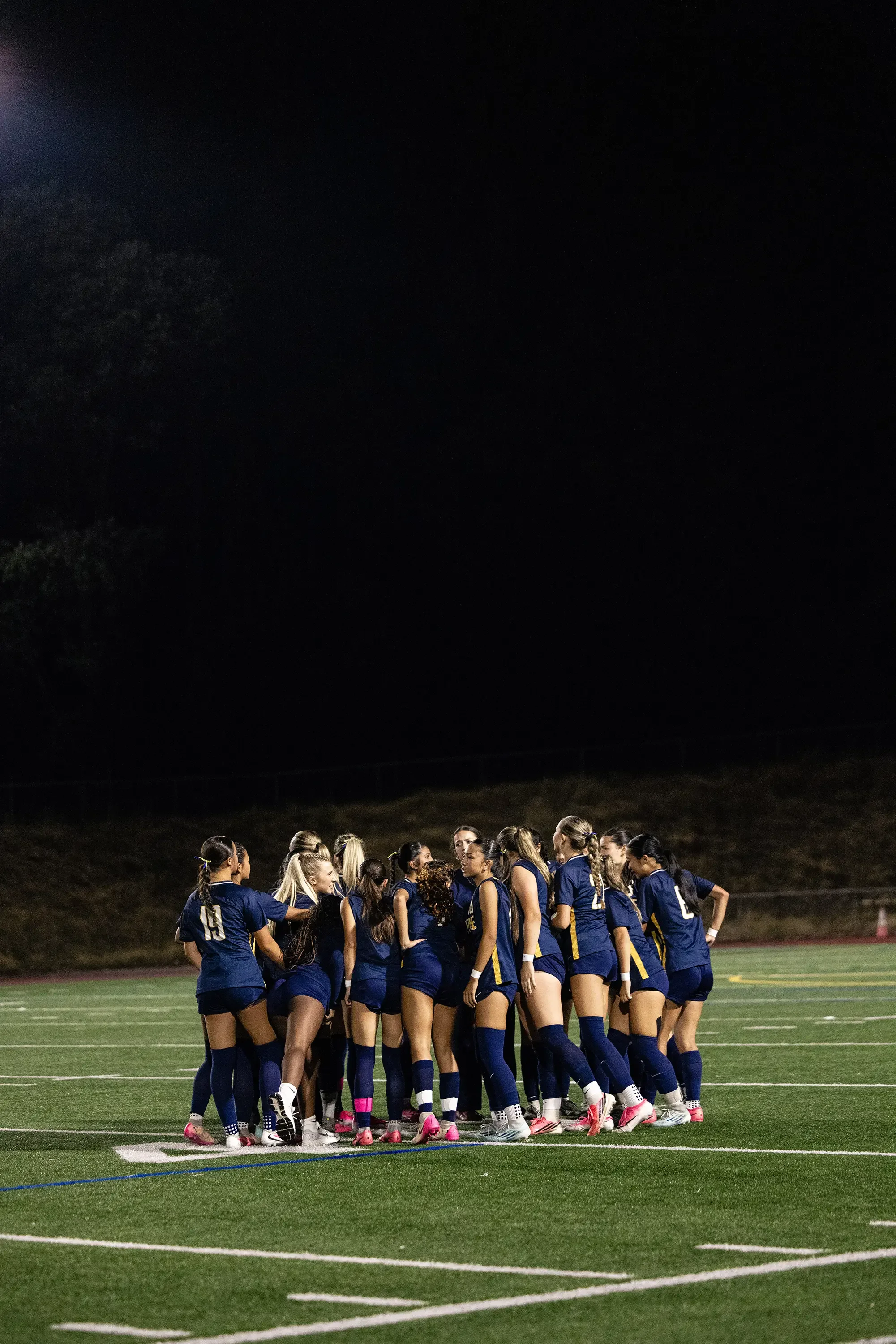 A group of female soccer players in blue uniforms huddled together on a well-lit outdoor field at night, possibly during a team meeting or pep talk.