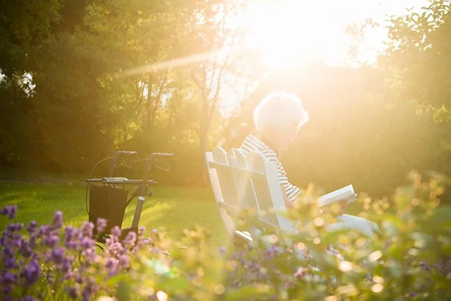 En eldre kvinne som leser en bok mens hun sitter i en hagemøbel omringet av blomster, med sollys som skinner gjennom trærne.
