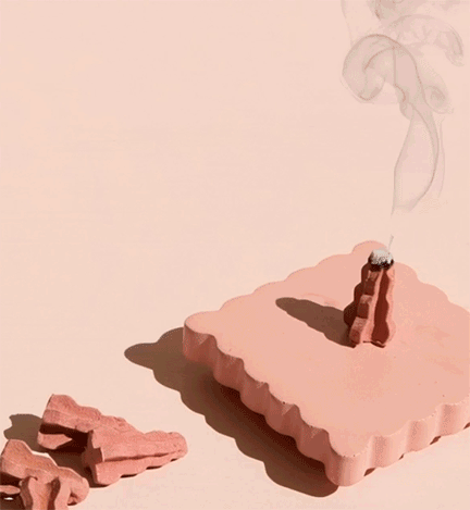 Pink soap with a small incense cone on top, producing smoke, and pieces of soap nearby on a pink surface.