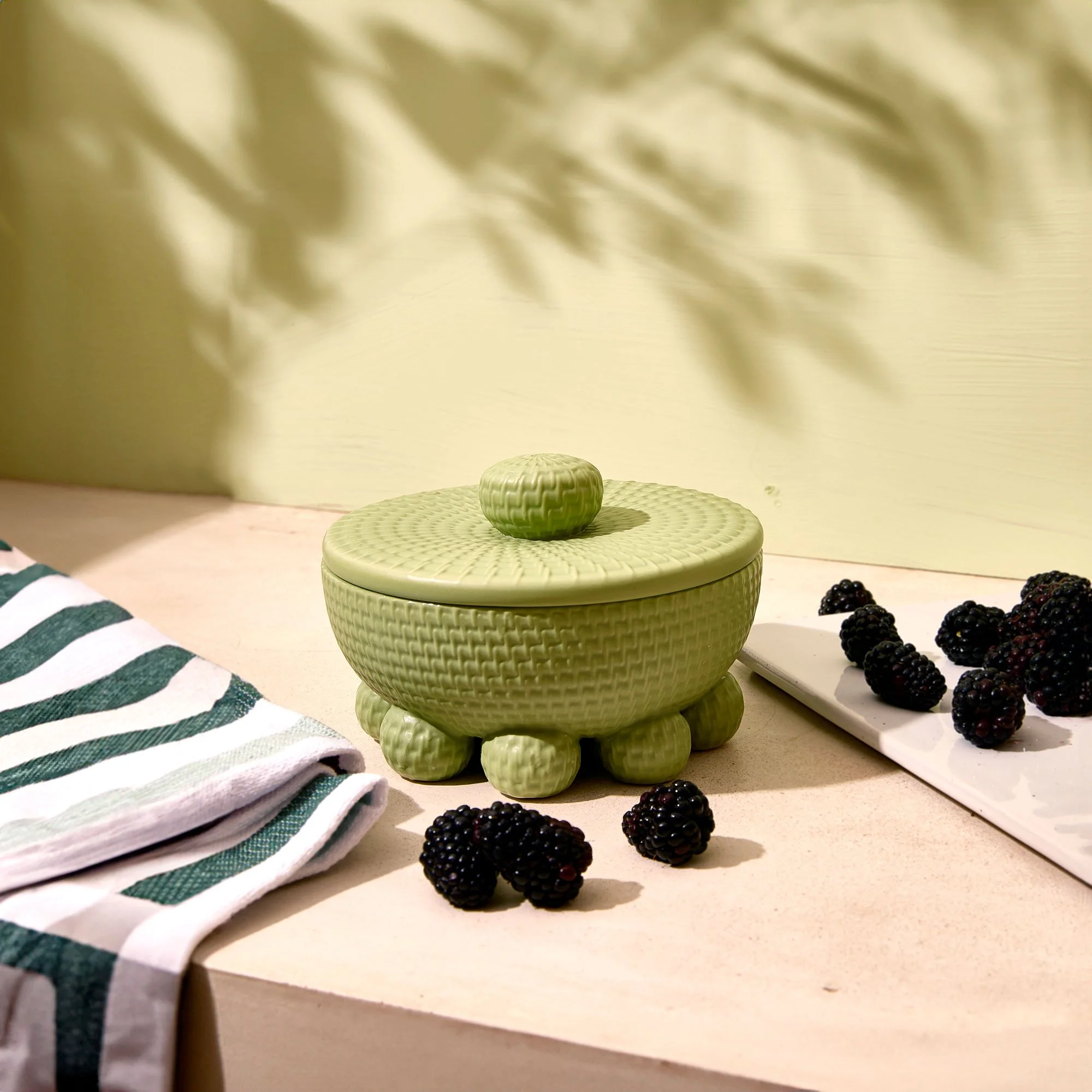 A green, textured, ceramic decorative box with a lid, placed on a beige surface, with blackberries scattered nearby and a striped green and white fabric partially visible in the foreground.