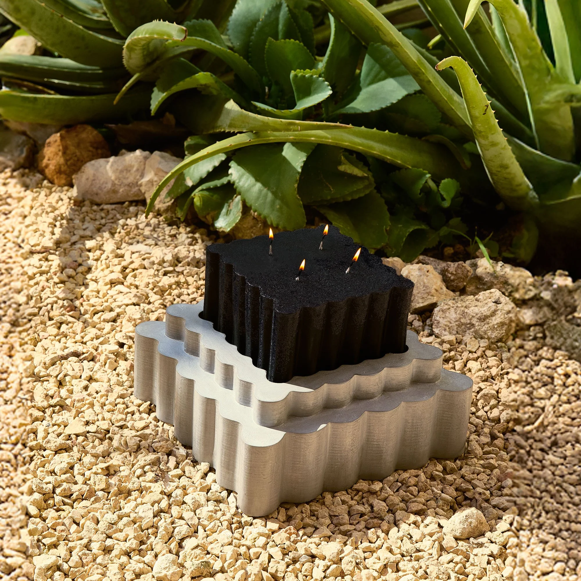 Close-up of a decorative candle holder with lit candles, placed on a bed of gravel surrounded by green succulent plants.