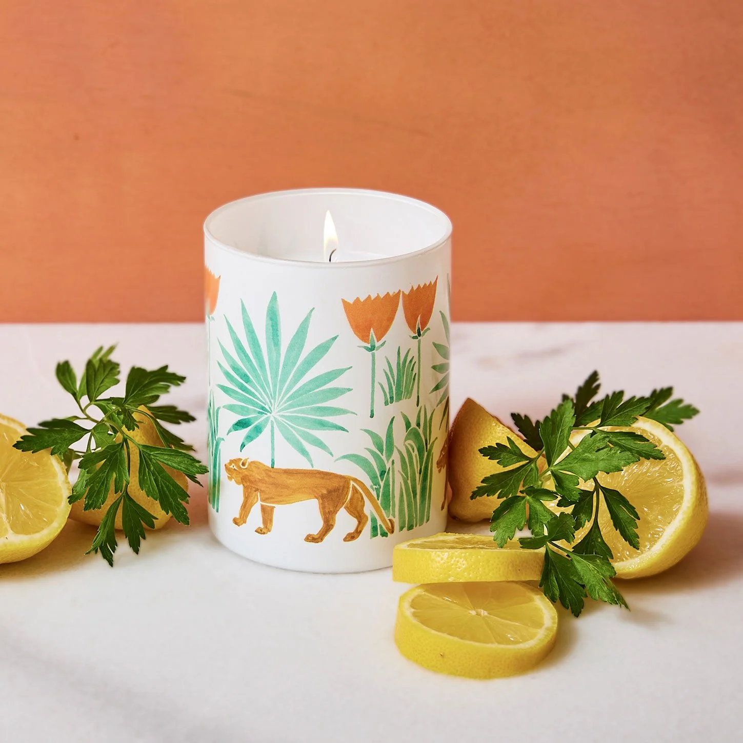 A white candle with jungle-themed illustrations, surrounded by lemon slices and green parsley sprigs on a white surface with a pinkish background.