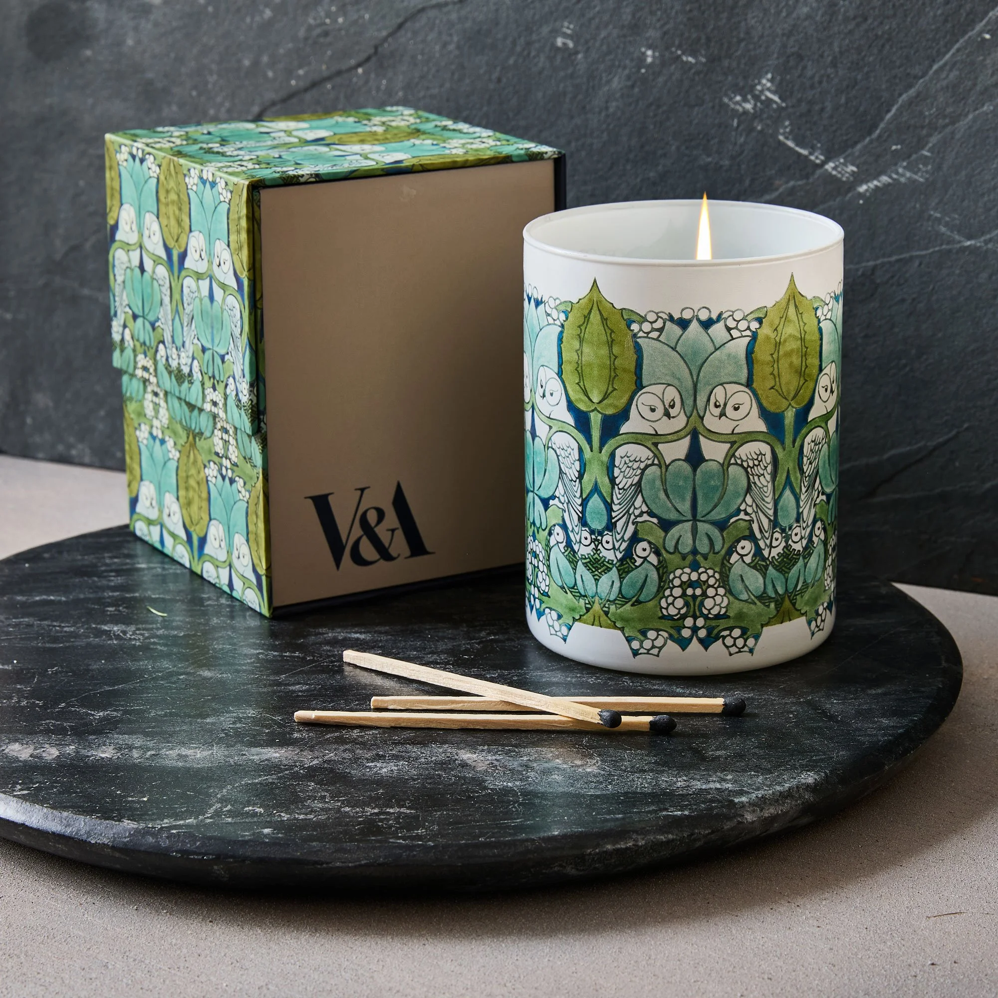 A lit candle with a decorative floral and leaf pattern in blue, green, and white, placed on a black slate surface alongside a matching box and two matchsticks.