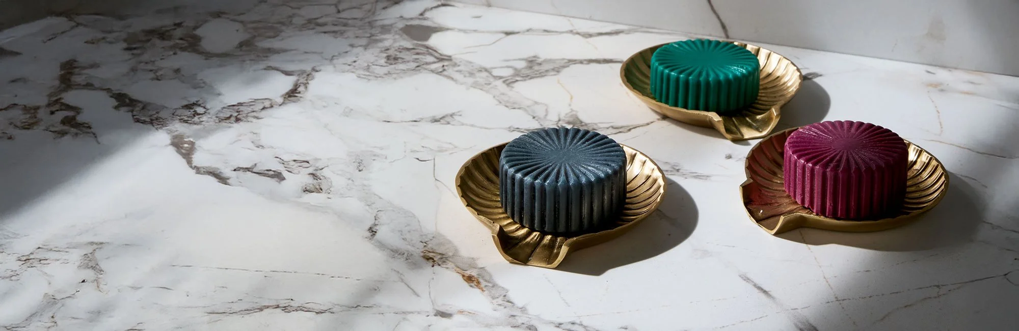Three colorful wax desserts in green, blue, and pink, each on a gold leaf-shaped plate, placed on a white marble surface.