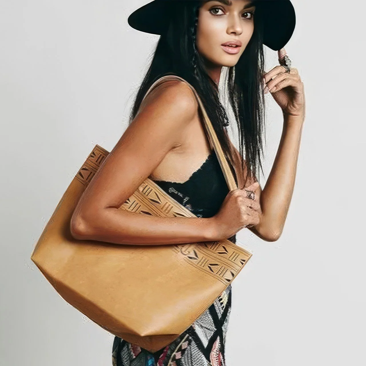Woman wearing a black wide-brim hat, black top, and patterned skirt, holding a carved wooden handbag against a plain light gray background.