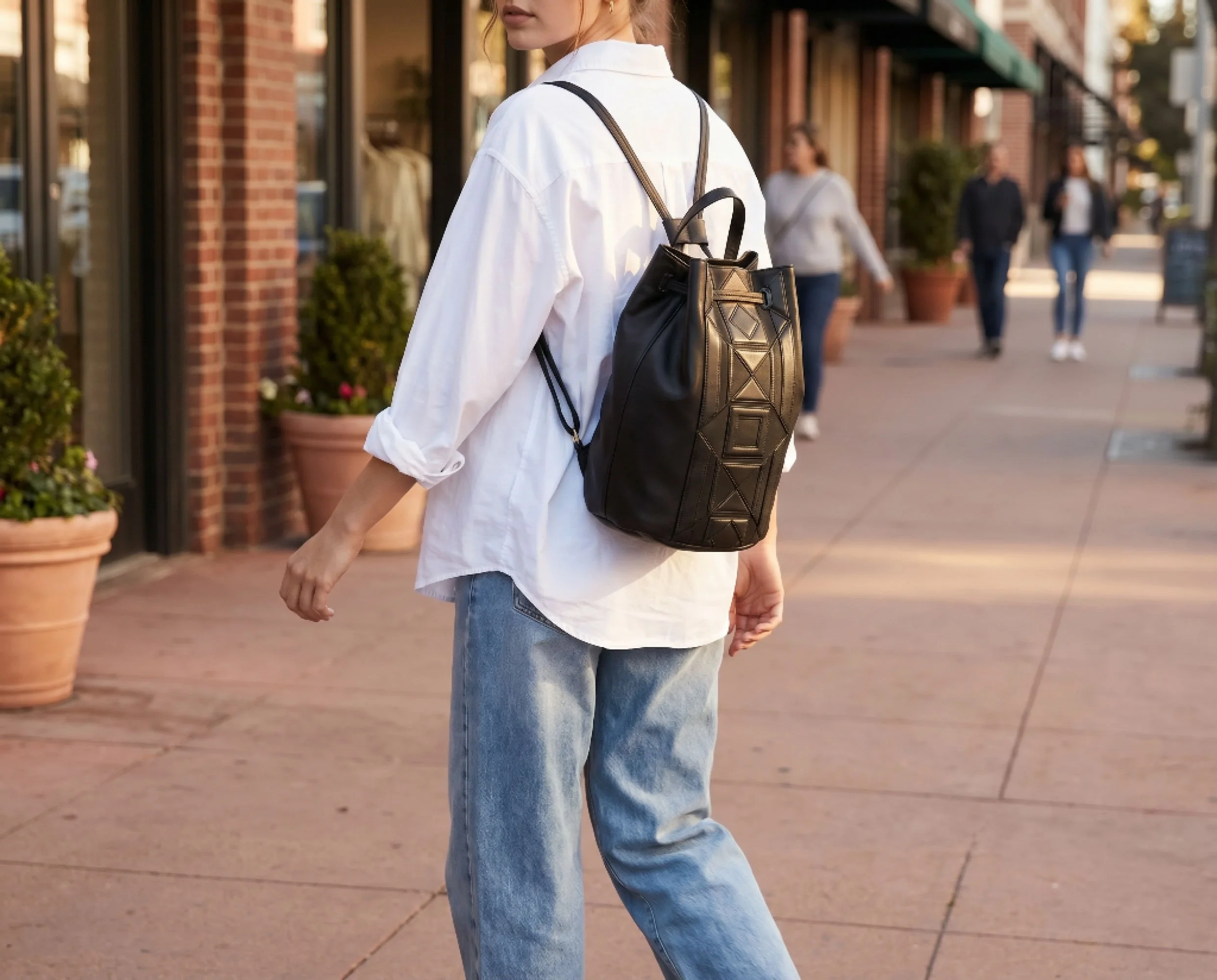 A woman wearing a white shirt and blue jeans standing outdoors in an urban shopping district with potted plants and storefronts, and other pedestrians walking in the background.