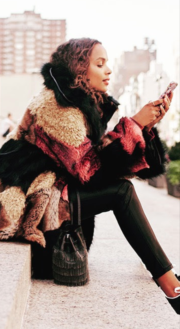 A woman in a colorful, patchwork fur coat sitting on a city sidewalk, looking at her phone.
