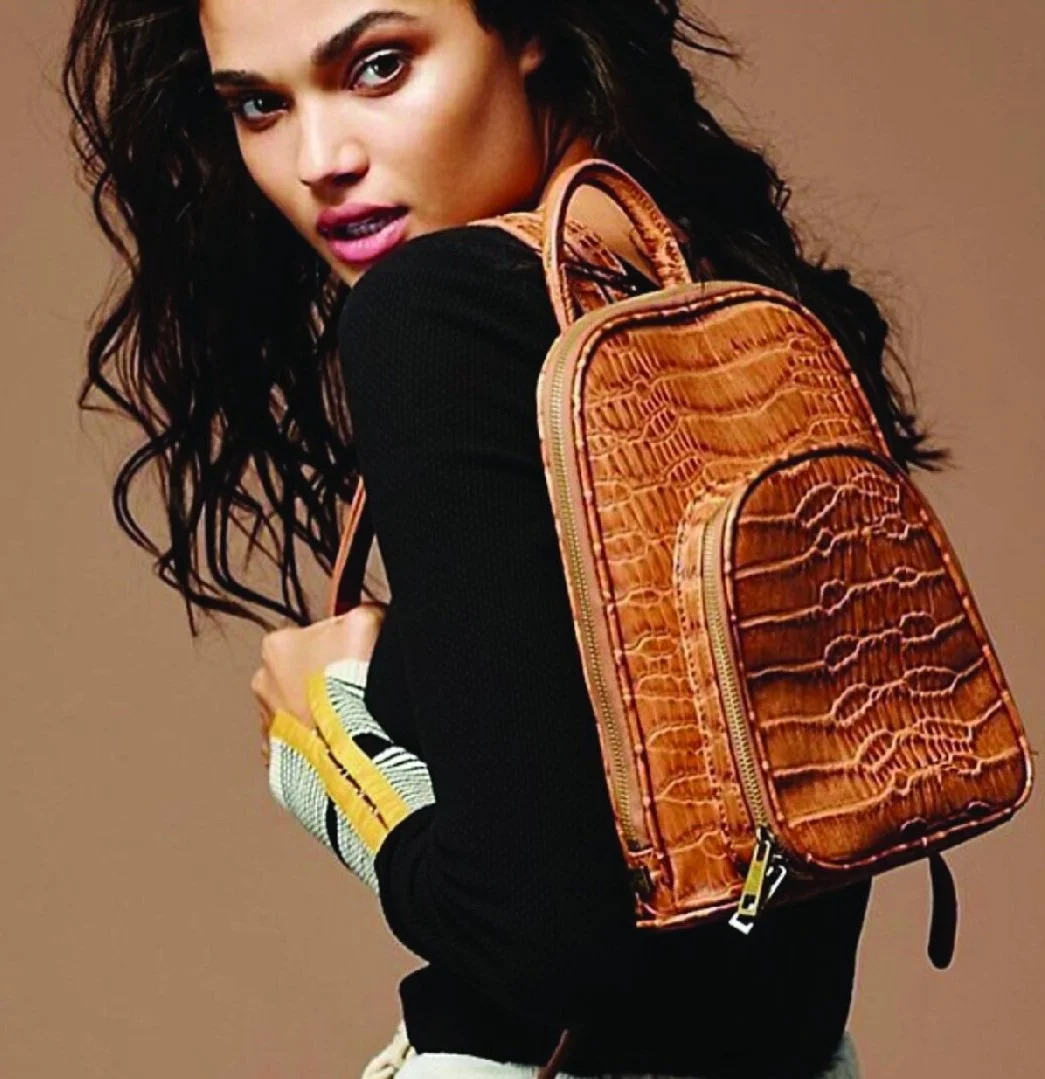 A woman with dark, wavy hair is wearing a black long-sleeve top and is carrying an orange, crocodile-patterned backpack over her shoulder. She is looking towards the camera with her lips slightly parted, showing a confident expression, against a plain tan background.