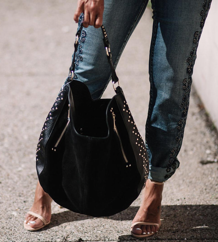 Person standing outdoors holding a large black handbag with studs and zippers, wearing distressed jeans and open-toe heels.