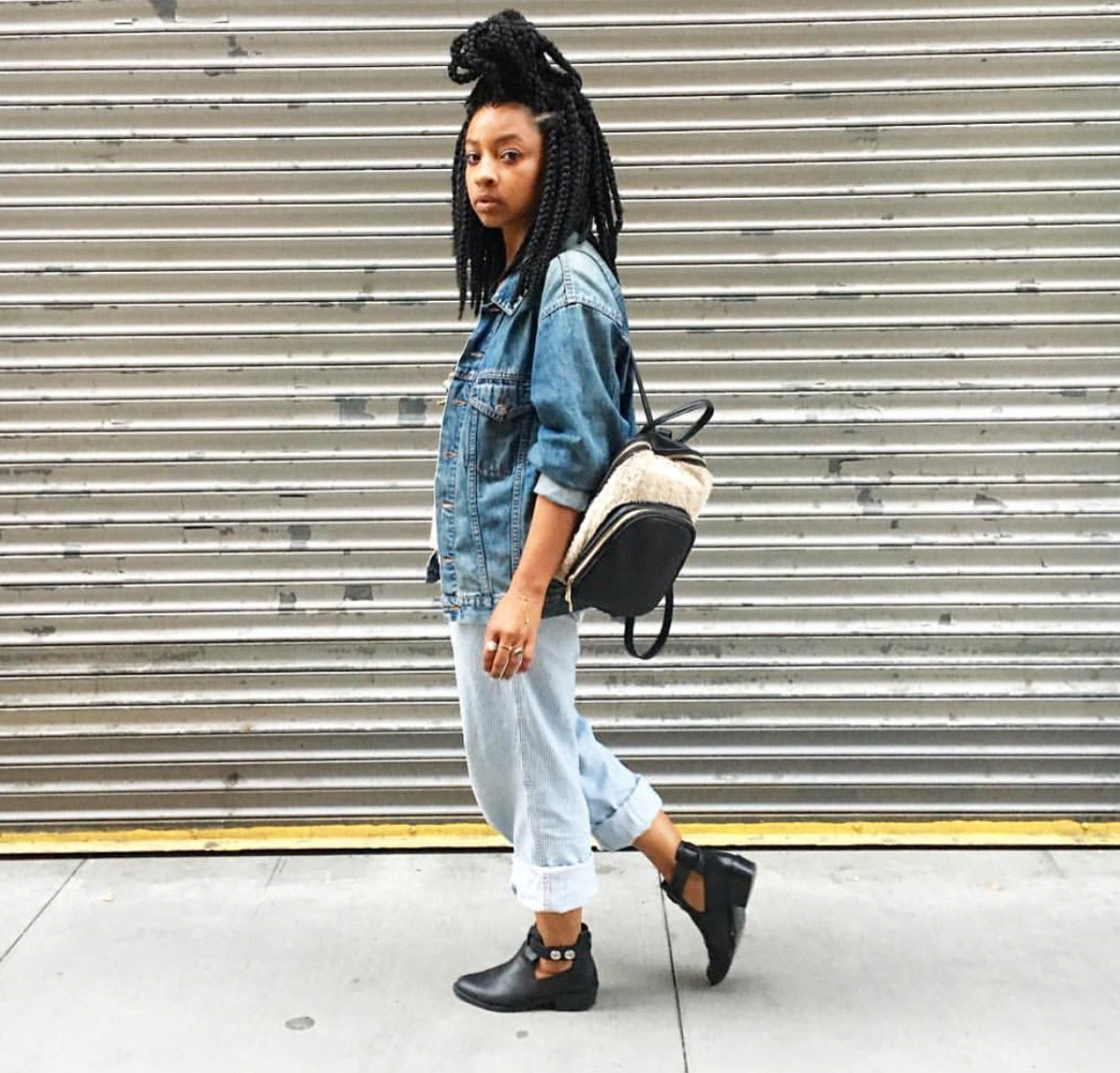 A woman with styled black braids wearing a denim jacket, light jeans, and black ankle boots, standing in front of a metal security shutter