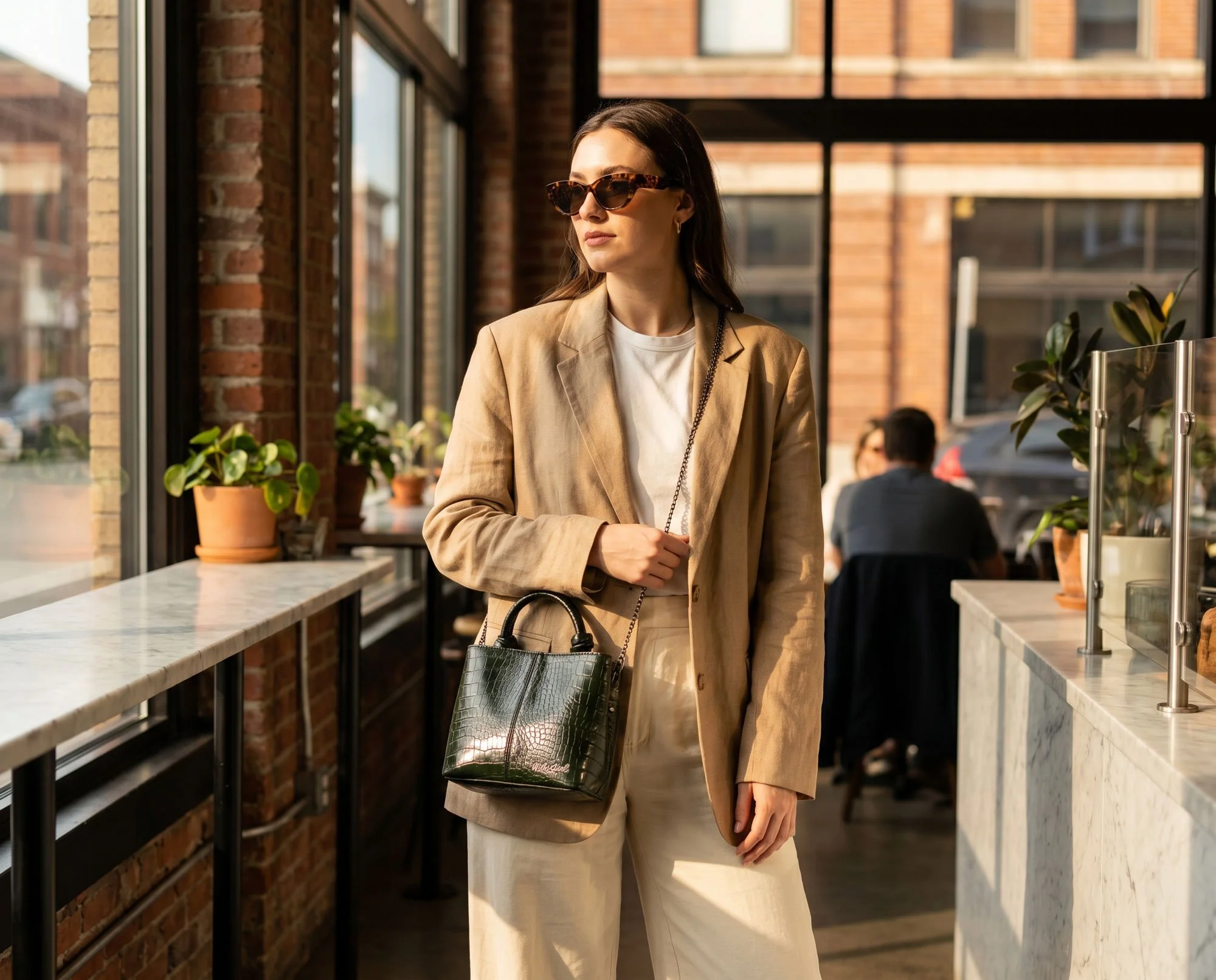 A woman with long dark hair wearing sunglasses, a beige blazer, and beige pants standing inside a modern building with large windows, brick walls, and potted plants, looking away from the camera.