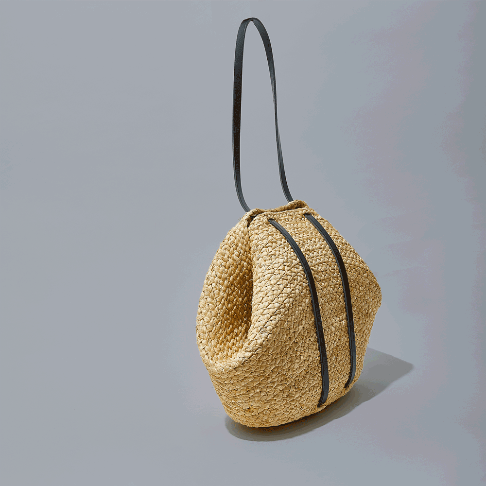 A woven straw handbag with black strap handles on a grey background.