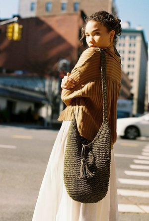 Young woman standing on city street with buildings in background, wearing a brown jacket and carrying a large gray woven bag.