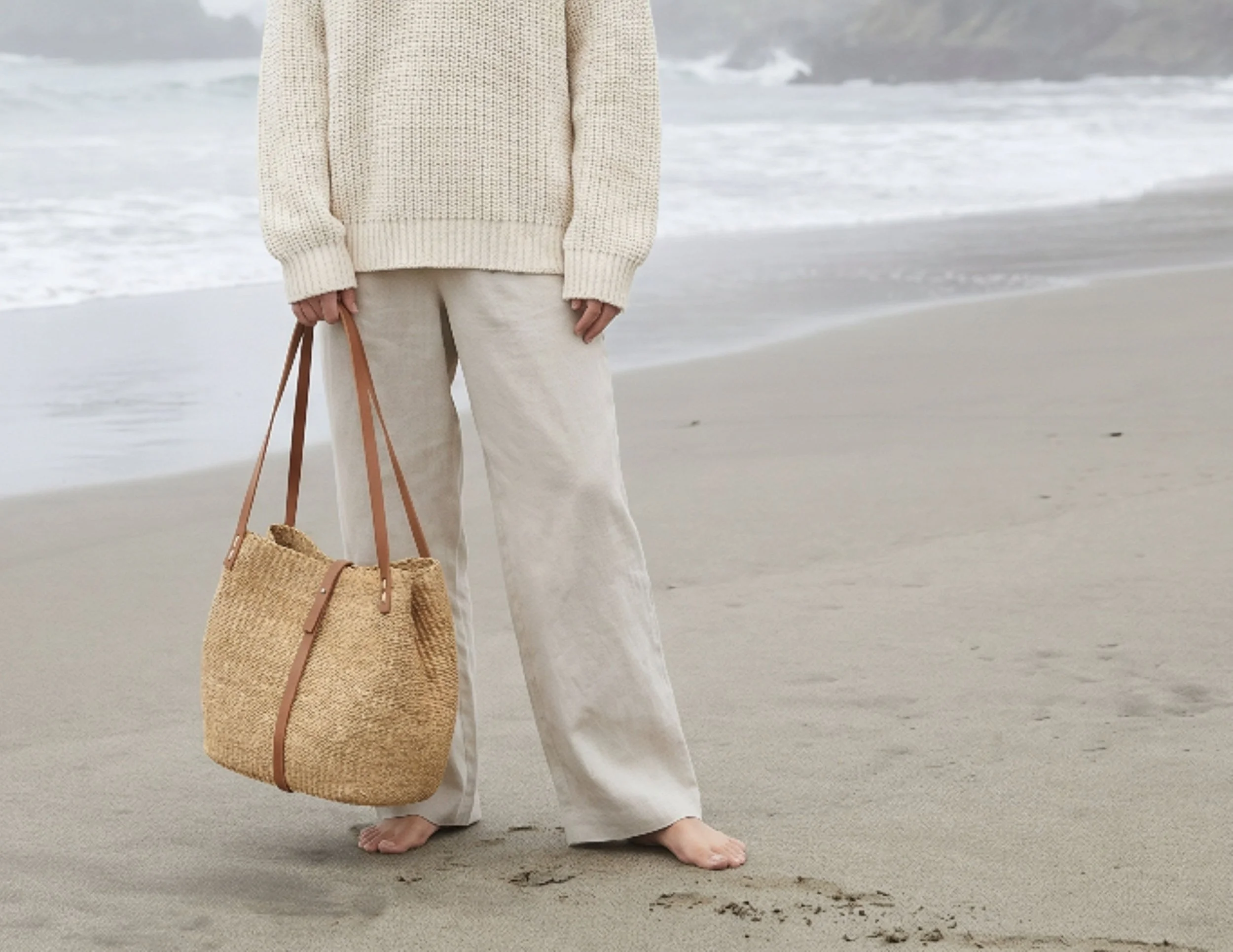 Person standing on the beach holding a woven tote bag with a tan strap, barefoot, wearing loose white pants and a cream-colored oversized sweater, with the ocean and cloudy sky in the background.