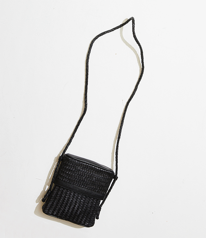 Black woven crossbody bag with a long strap hanging on a white surface.