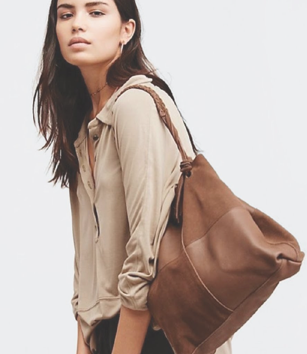 Young woman with dark hair wearing a beige shirt, carrying a large brown leather bag over her shoulder, standing against a plain background.