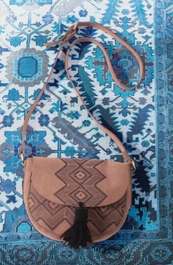 A tan shoulder bag with a geometric pattern and a black tassel, placed on a blue and white patterned rug.