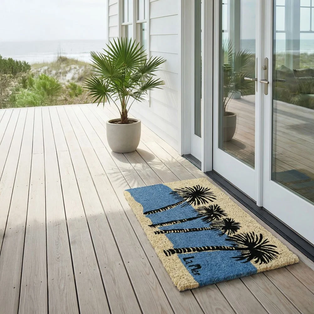 A wooden deck with a potted palm tree, sliding glass door, and a palm tree themed outdoor rug. The background shows sand dunes and greenery.