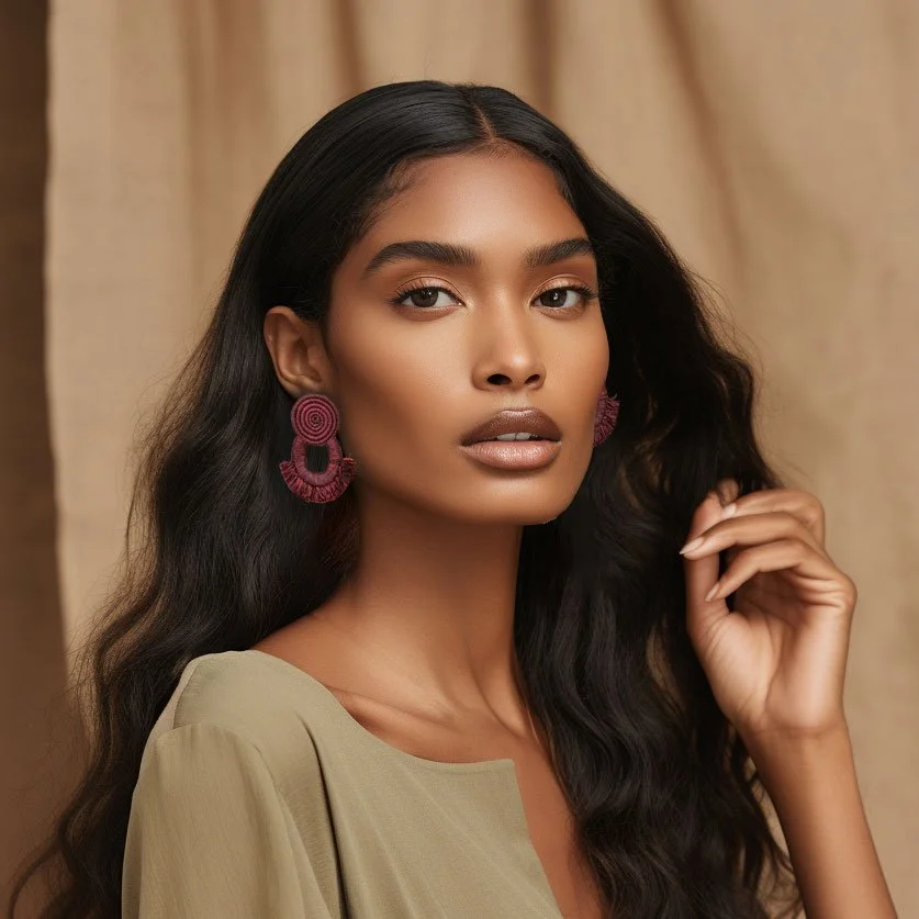 A woman with long wavy black hair and medium skin tone wearing mauve statement earrings and a beige top, posing indoors against beige curtains.
