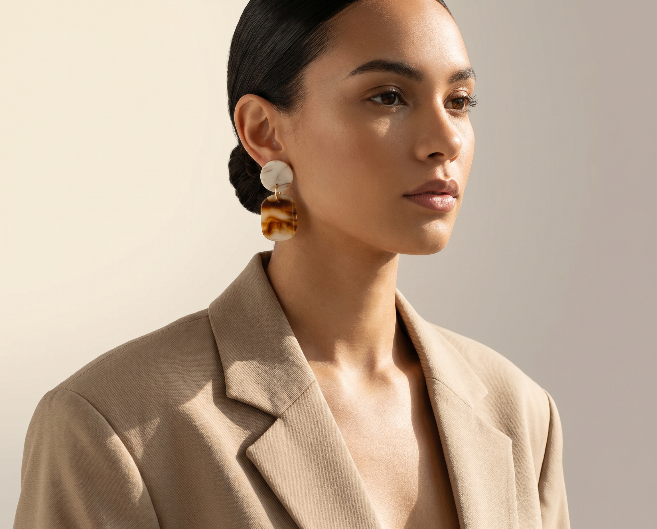 A woman with dark hair styled in a sleek bun, wearing large dangling earrings, a beige blazer, and a neutral makeup look, against a plain beige background.