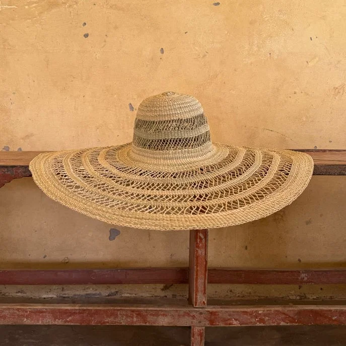 A large woven straw sun hat with a wide brim and a circular crown, resting on a rustic wooden surface against a beige wall.