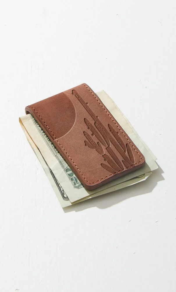 A brown leather card holder with a cactus design, holding some folded US dollar bills, placed on a white surface.
