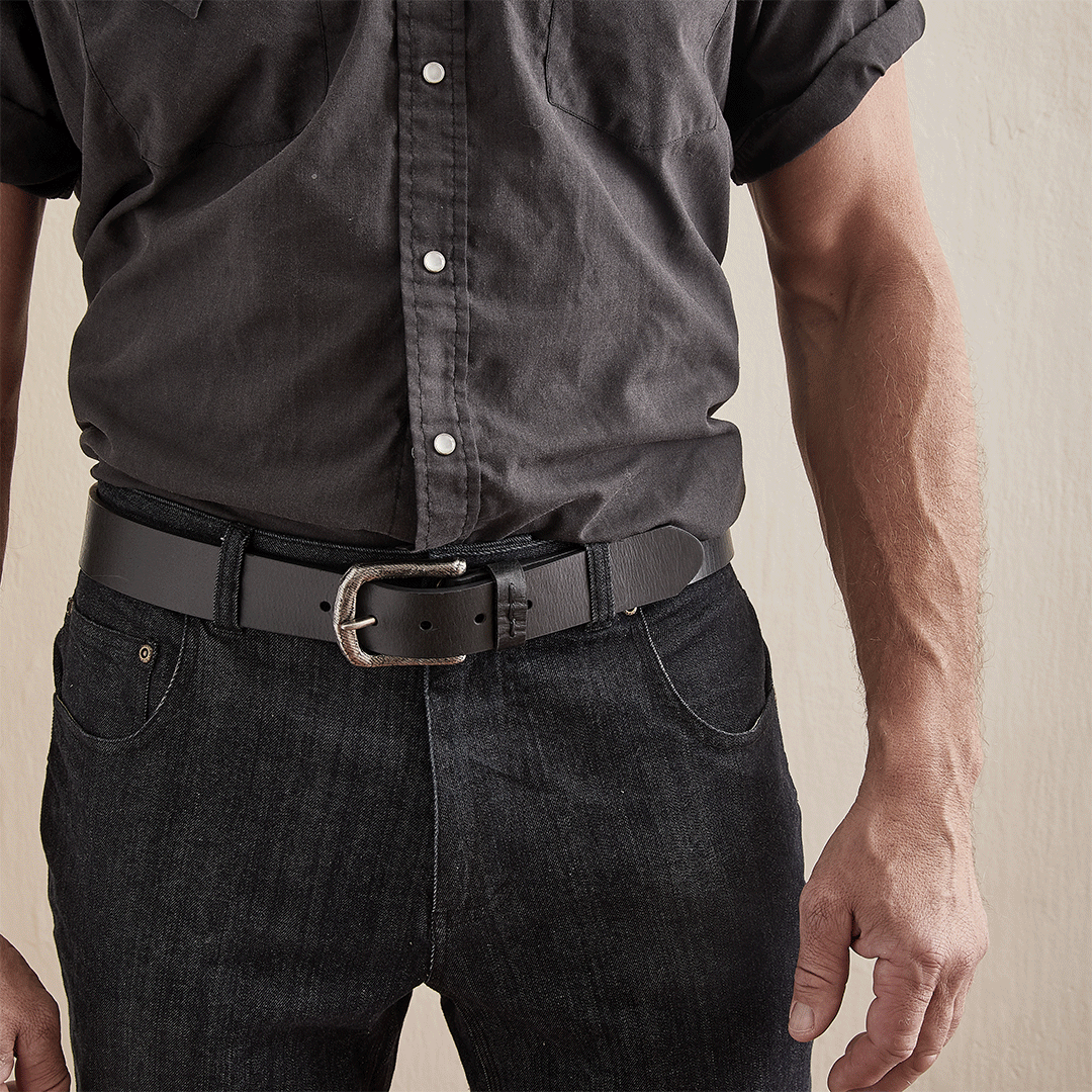 Close-up of a man wearing a black button-up shirt, black jeans, and a black belt, standing against a light-colored wall.