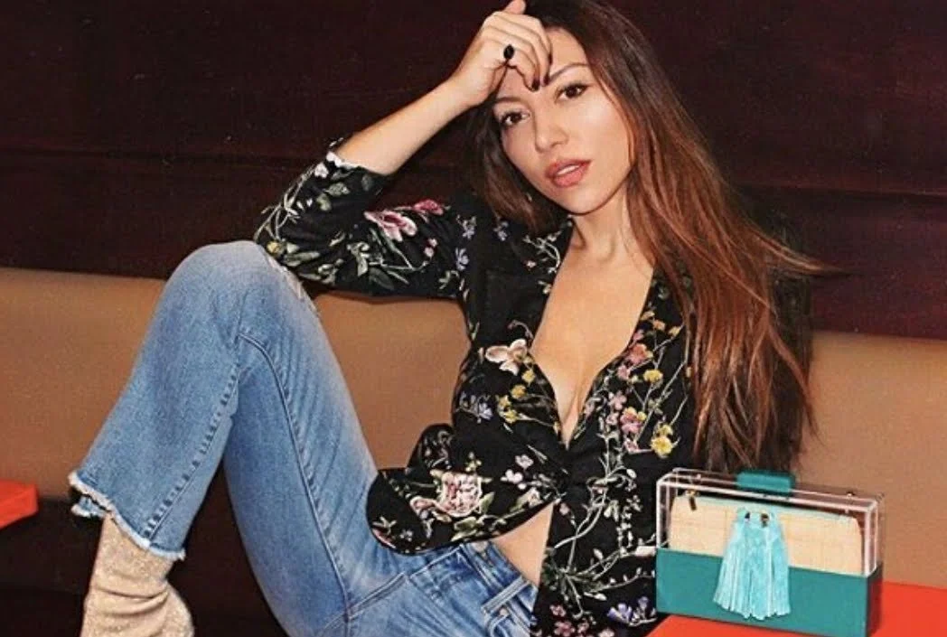 A young woman with long brown hair sitting on a bench with one knee up, wearing a floral black blouse, jeans, and beige socks.