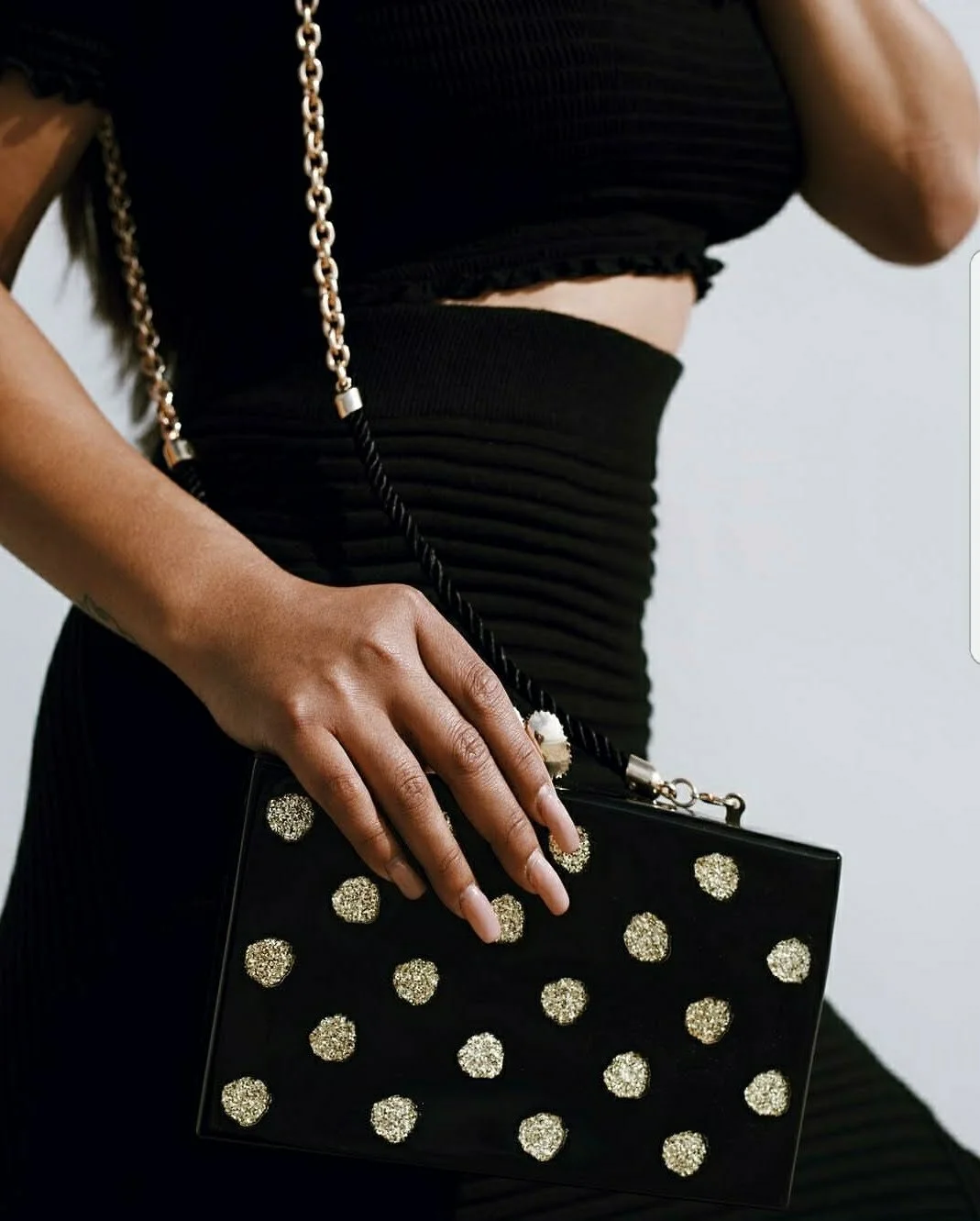 Person holding a black clutch purse with gold glittery polka dots, wearing a black crop top and skirt, with a gold chain and black strap handbag.