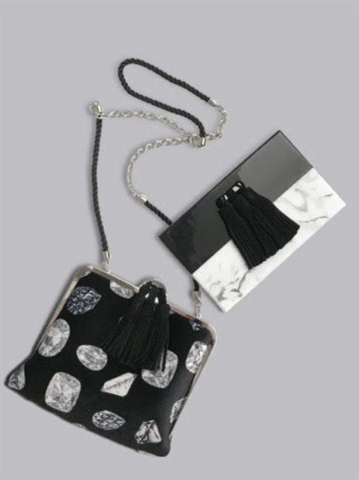Two small handbags with chain straps, one black with a diamond pattern and the other with a marble design, along with a black and white box containing a black tassel keychain, all placed on a gray surface.