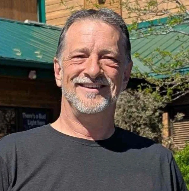 A smiling middle-aged man with gray hair and beard, wearing a black T-shirt, standing outdoors in front of a building with a green roof and some trees.