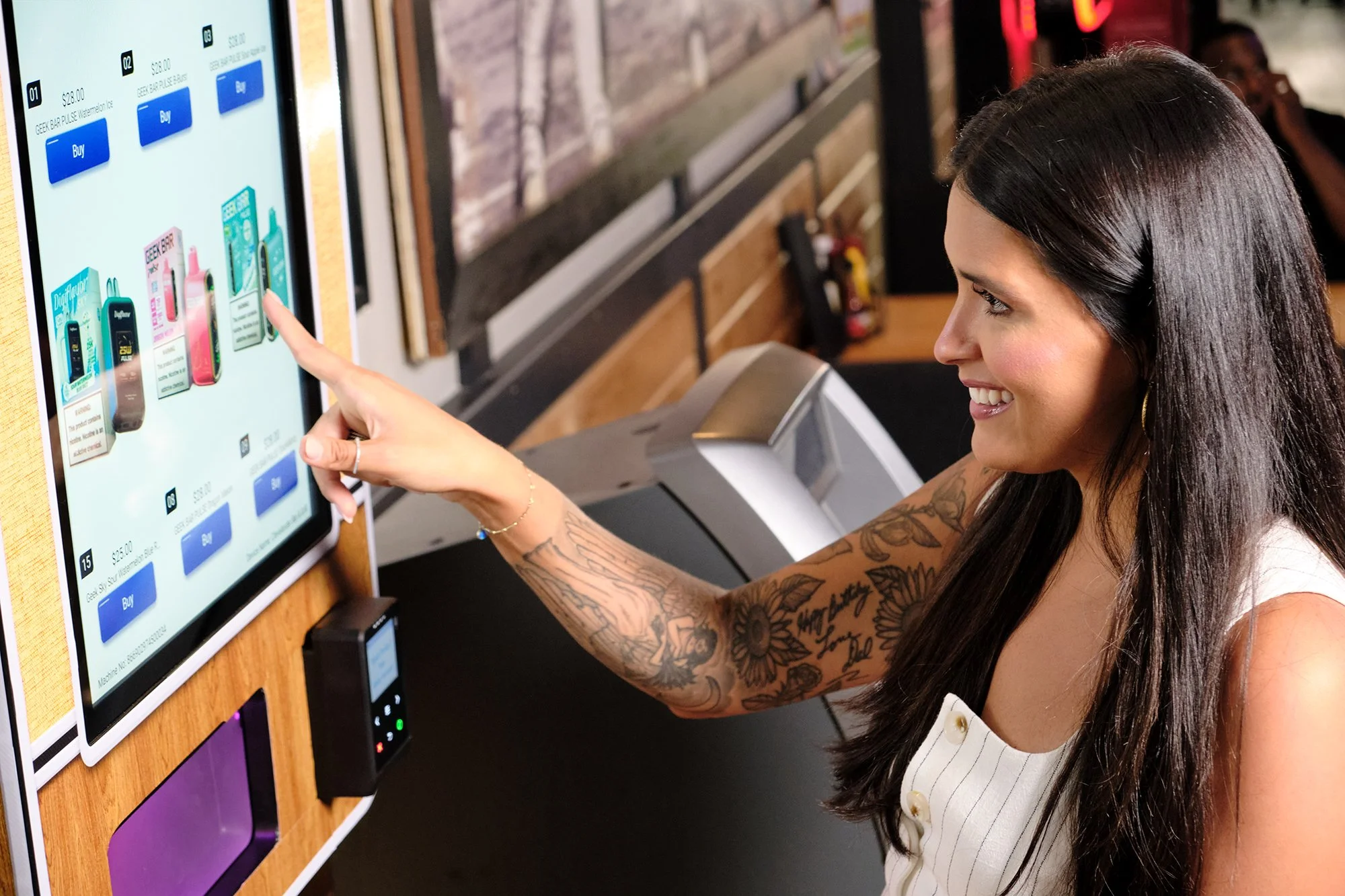A woman with long dark hair and tattoos on her arm using a touchscreen kiosk to purchase items such as a digital vaporizer or electronic cigarette, smiling as she taps on the screen in a retail setting.