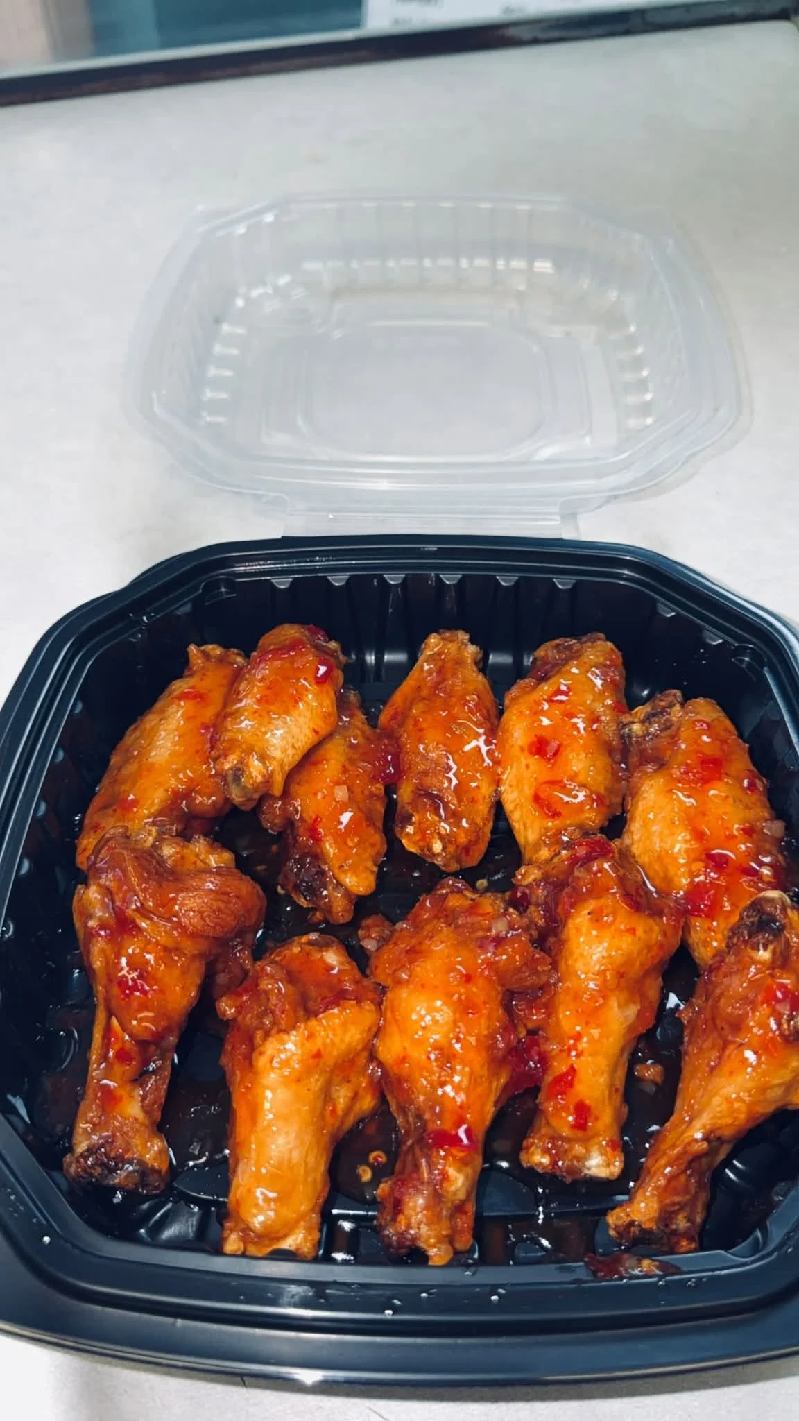 Tray of spicy glazed chicken wings with a reddish sauce on top, inside a black plastic container with a clear lid.
