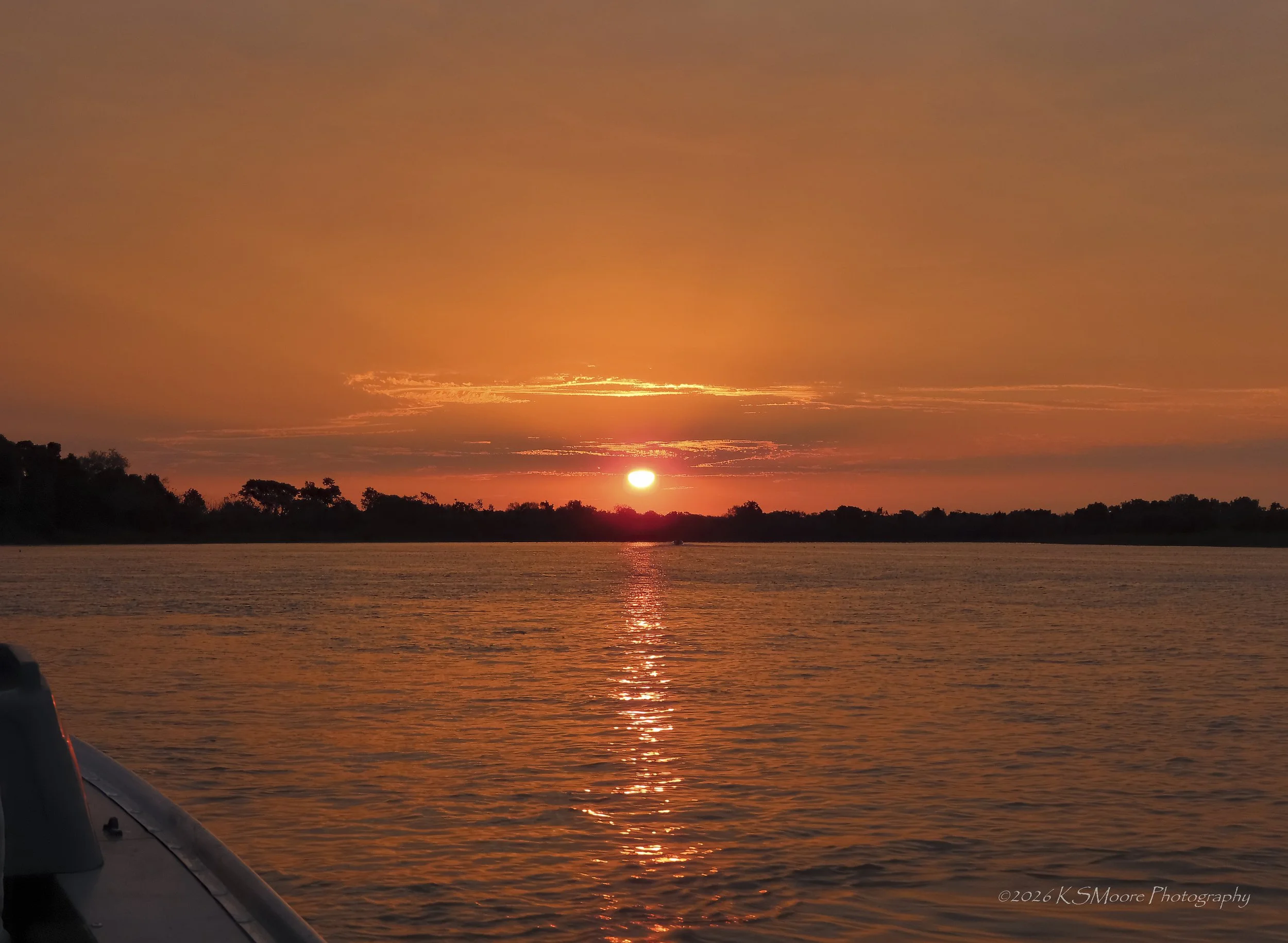 Sunset over a body of water with orange and pink hues, silhouettes of trees on the horizon, and a small boat visible in the distance.